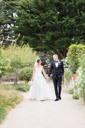 Bride and groom walk hand in hand along a garden path, she in a white gown, he in a black tuxedo.