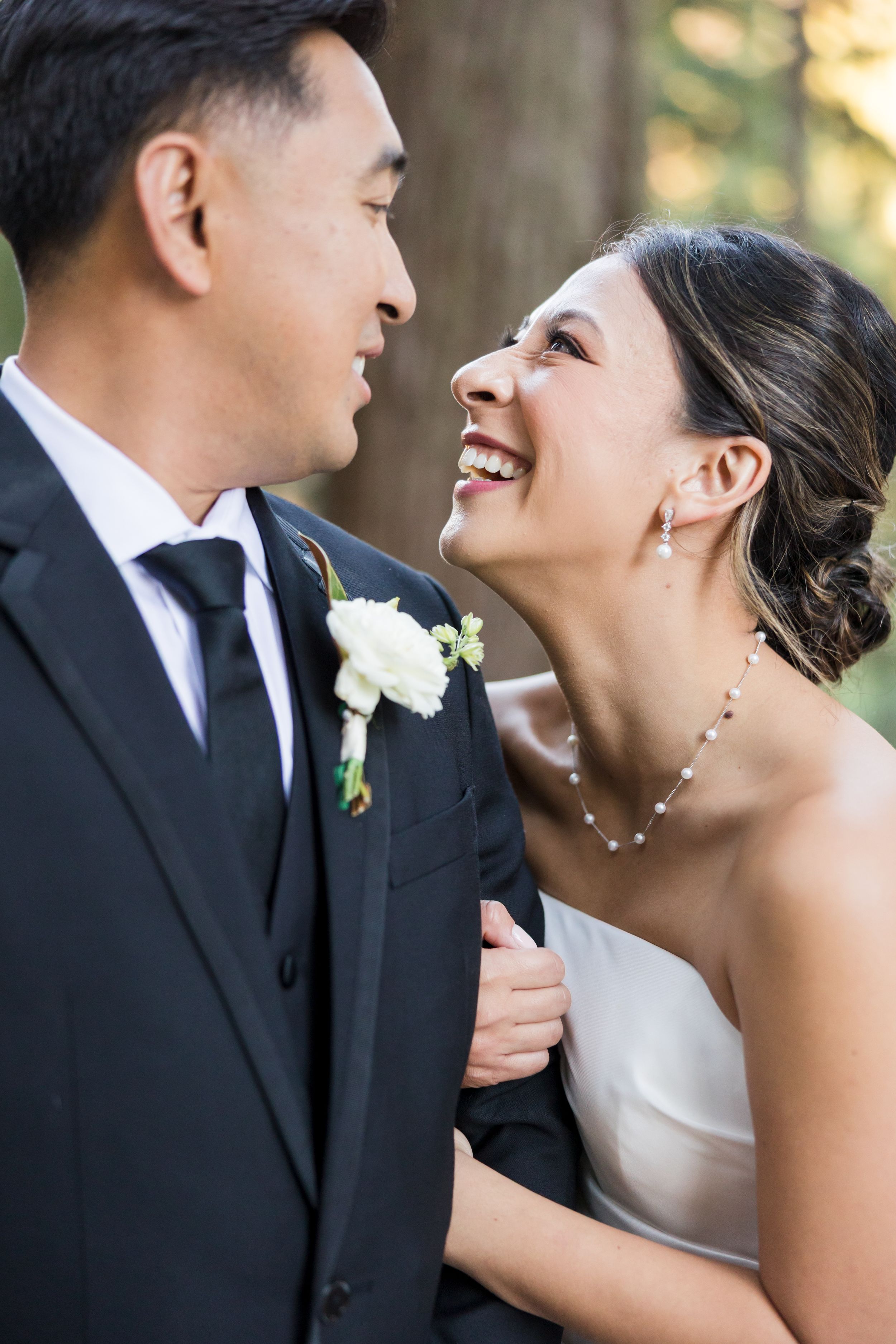 Joyful bride in white dress laughs with groom in black suit and boutonniere during outdoor wedding portrait.