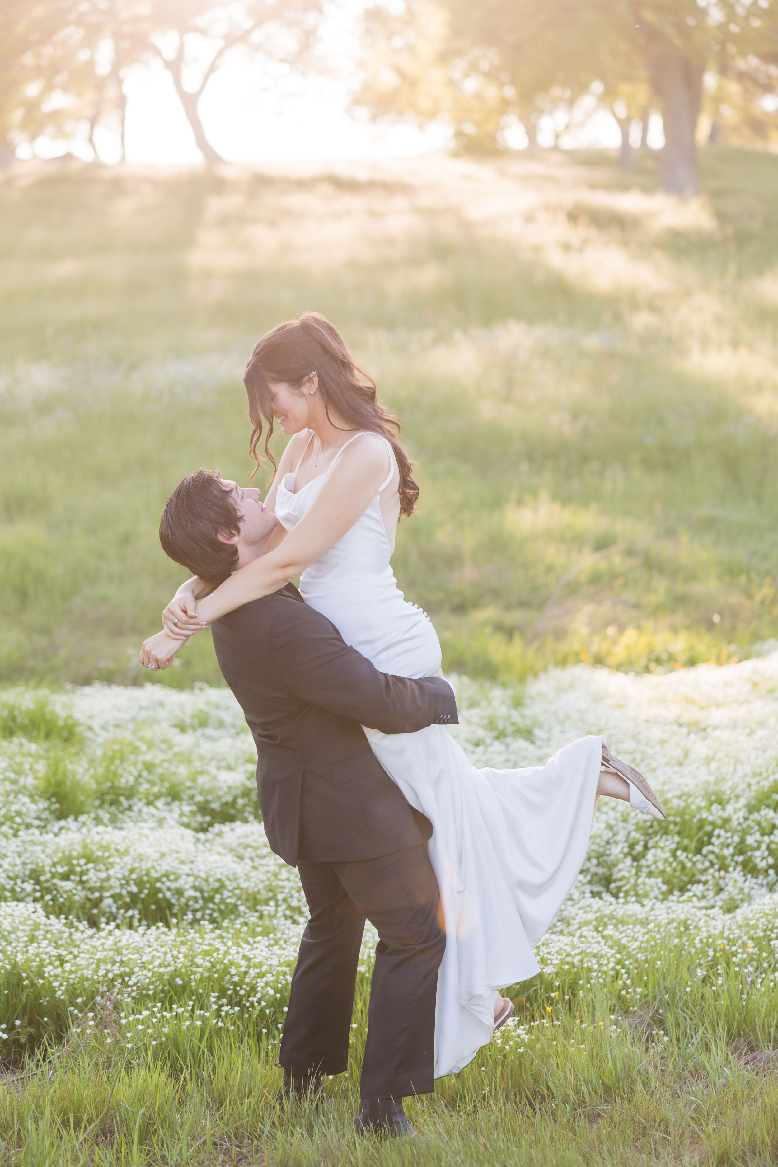Groom lifting bride in white dress in sunlit meadow with wildflowers during romantic outdoor wedding photo session.