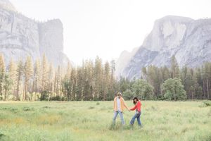 Couple holding hands walking through a green meadow in Yosemite Valley with granite cliffs and pine trees in background.