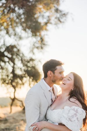 A bride and groom share a romantic golden hour moment outdoors, with the groom kissing her cheek as she laughs joyfully.