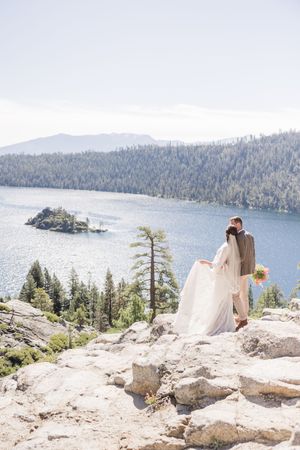 Bride and groom stand on rocky overlook above Emerald Bay at Lake Tahoe, surrounded by pine forests and mountains.