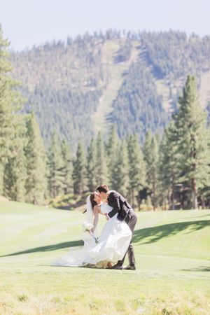 Bride and groom share a romantic dip kiss on a golf course with pine trees and mountain backdrop.