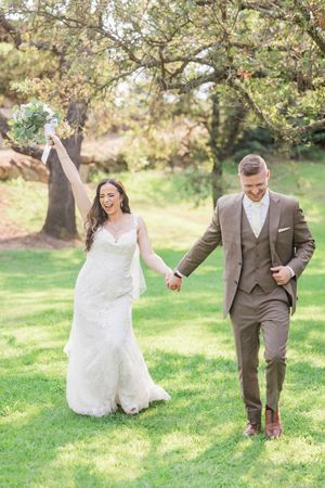 Joyful bride in white lace gown raises bouquet while groom in brown suit holds her hand on green lawn.