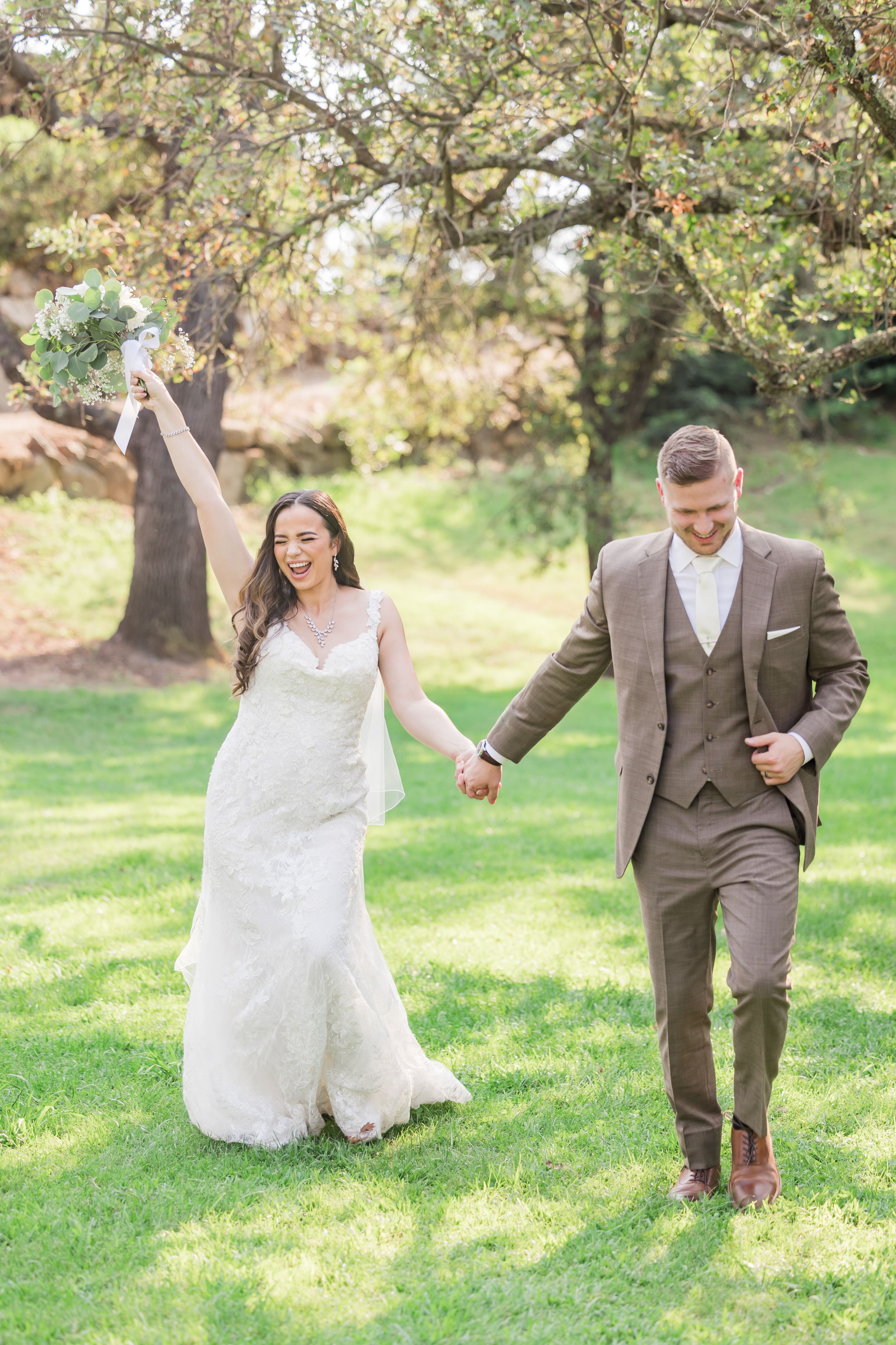 Joyful bride in white lace gown raises bouquet while groom in brown suit holds her hand on green lawn.