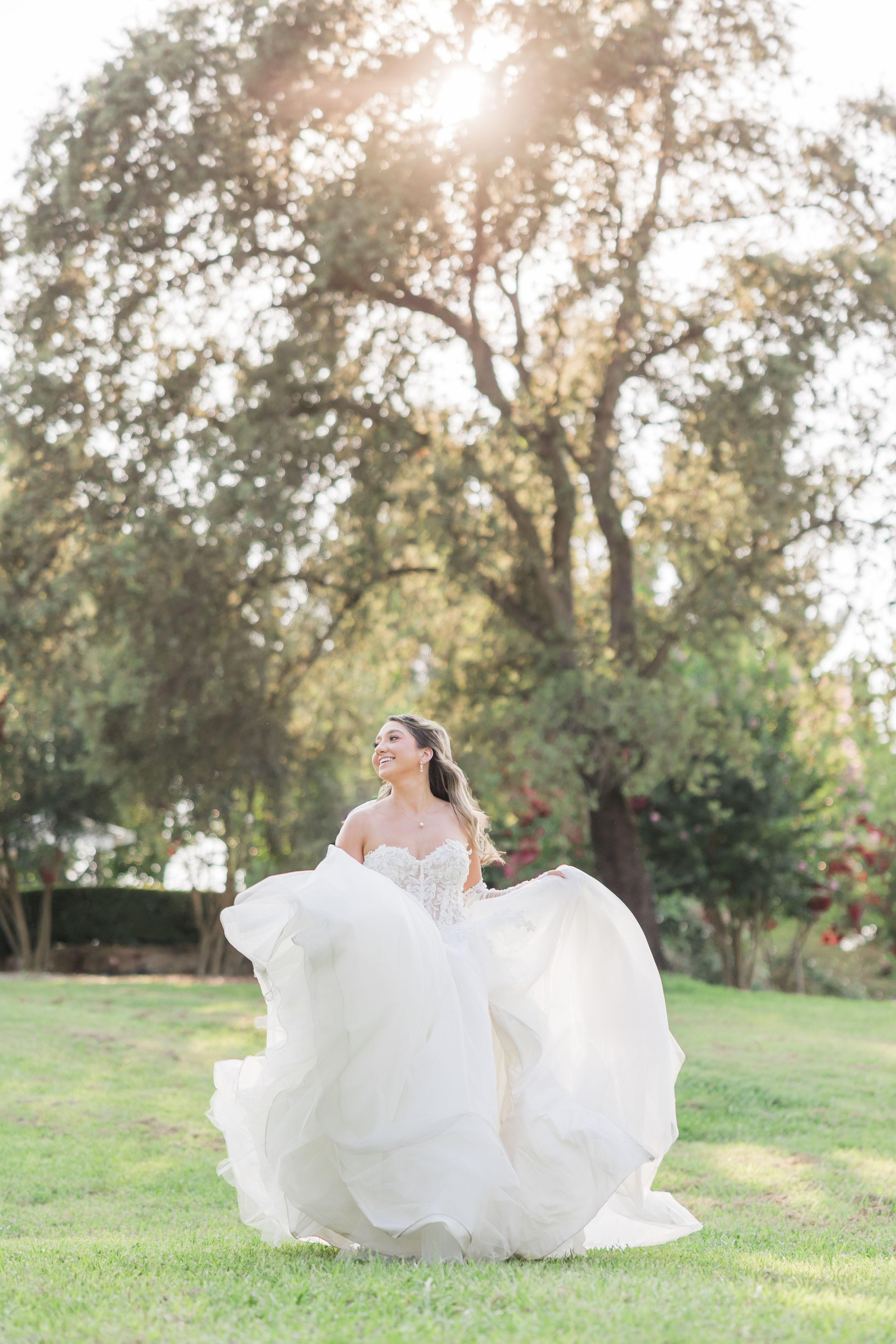 Bride in flowing white gown twirling outdoors on green lawn with sunlit trees in background.