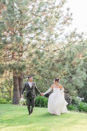 Bride and groom holding hands walking on green grass with tall pine trees in background at outdoor wedding.
