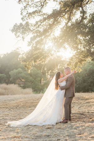 Bride and groom embrace at golden hour under oak trees in a sunlit field, her long veil trailing behind.