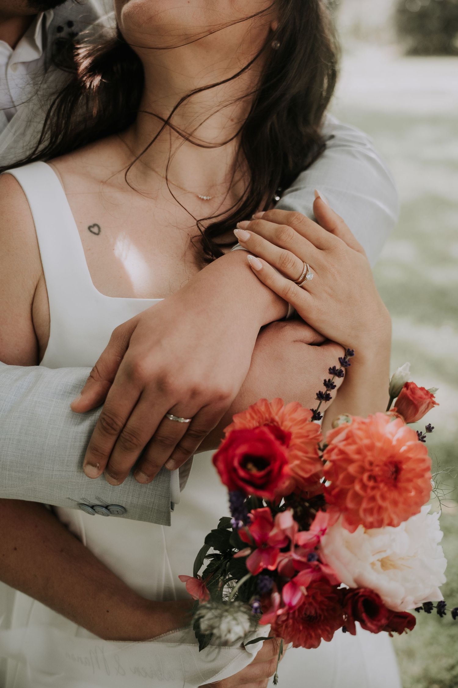 Bride in white dress holding colorful bouquet embraced by groom, both wearing wedding rings, outdoor wedding photo.