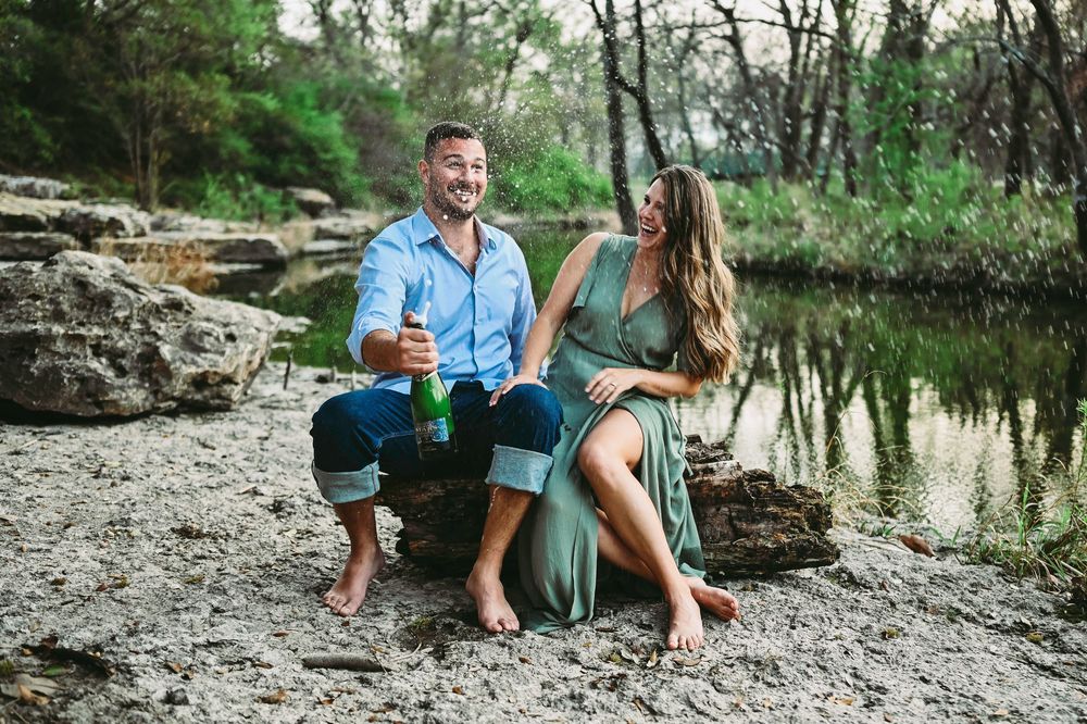 Couple sitting on rocky riverbank sharing drinks and laughing together outdoors.