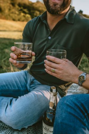 Couple holding whiskey glasses outdoors on a blanket during a golden hour picnic.