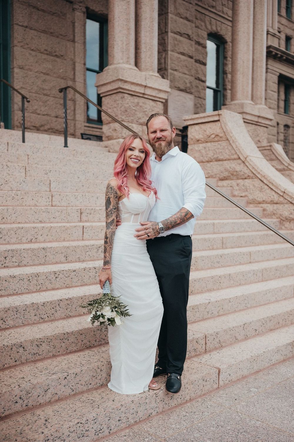 Tattooed couple in wedding attire poses on stone steps of a grand building, bride holding a bouquet.