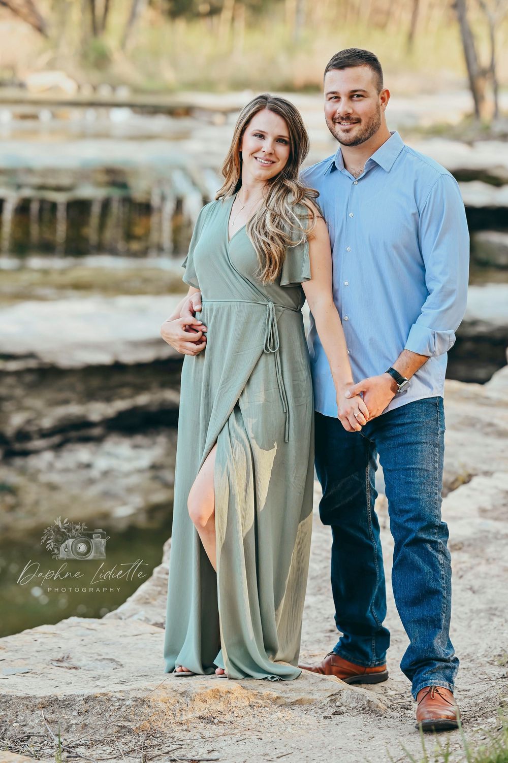 Smiling couple standing on rocks near a waterfall, dressed in elegant casual attire.