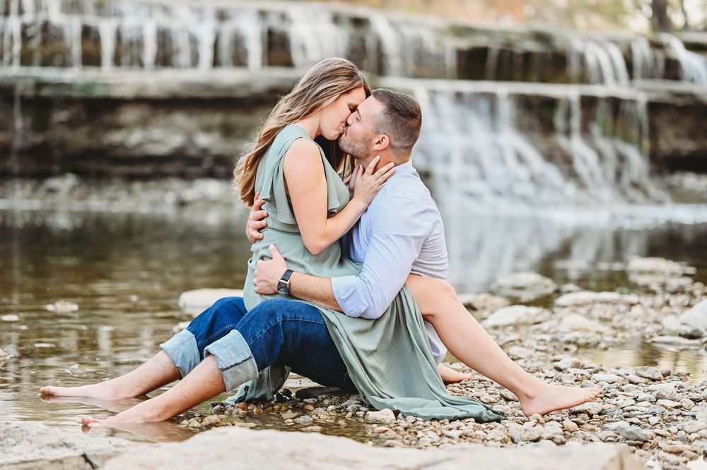 Couple sharing a kiss while sitting on rocky riverbank in front of a scenic waterfall.