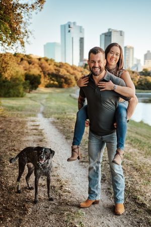 Smiling couple with a dog poses on a park trail with city skyline in the background.