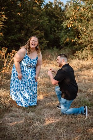 Man proposes on one knee to laughing woman in blue floral dress in a golden field.
