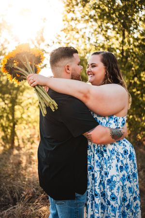 Joyful couple embraces holding sunflowers in a sun-drenched outdoor engagement photo session.