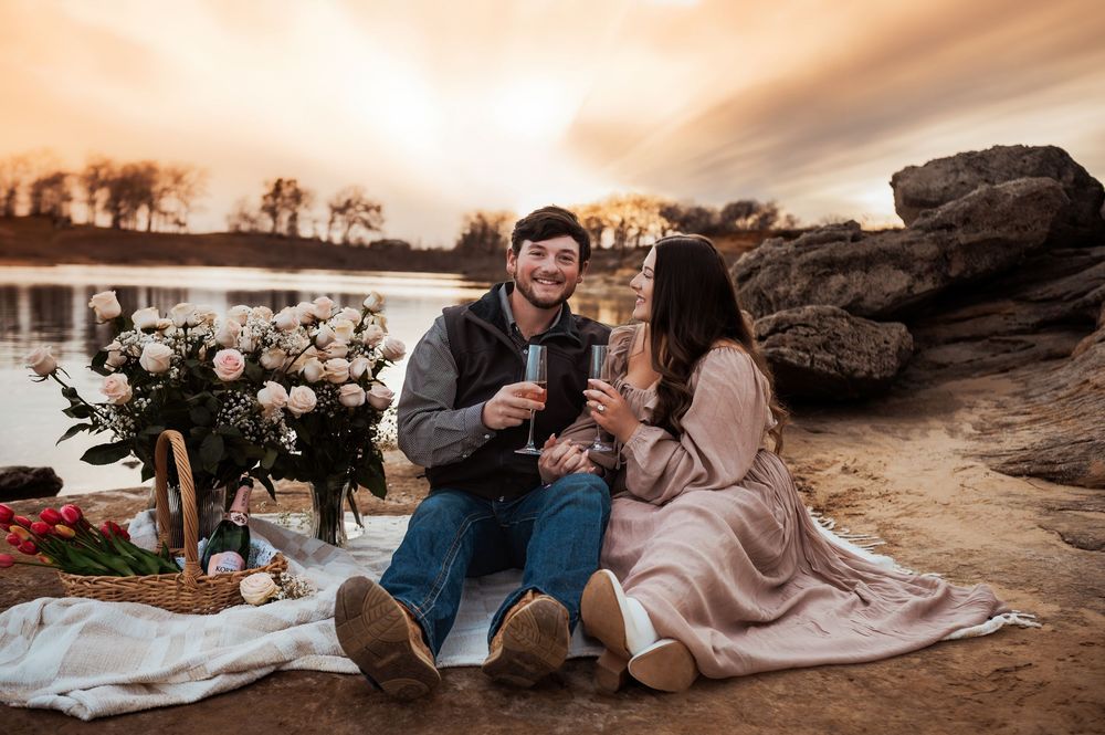 Engaged couple enjoys a romantic lakeside picnic at sunset with flowers and champagne.