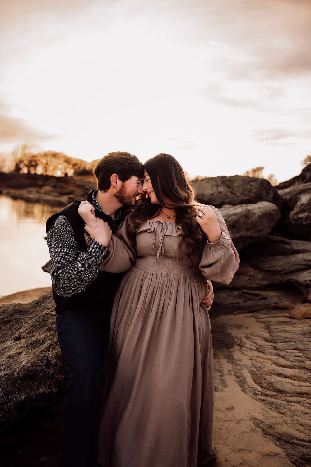 Couple sharing a romantic moment on rocky shore at sunset, woman in flowing mauve dress, man kissing her cheek.