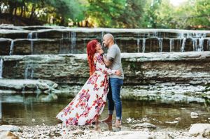 Couple embracing near a waterfall, woman in floral maxi dress with red hair, standing in shallow water.