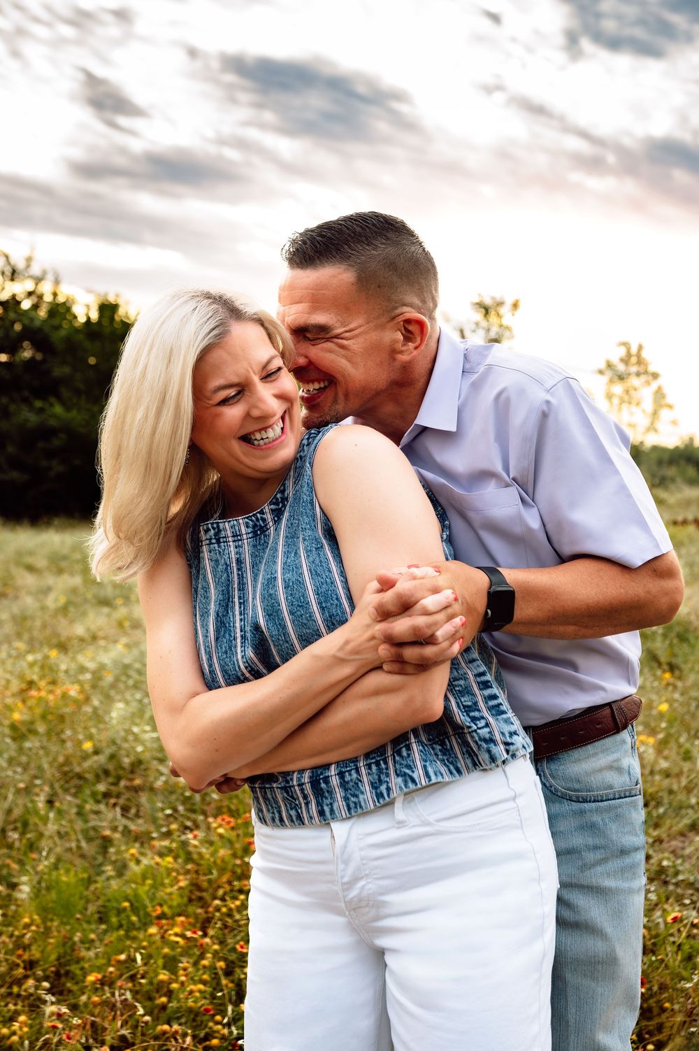 Middle-aged couple laughing joyfully in a golden field at sunset, woman in blue top and white pants.