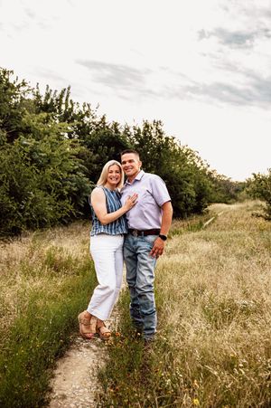 Smiling couple posing on a nature trail surrounded by tall grass and green trees under cloudy sky.