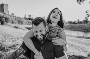 Black and white photo of smiling man giving laughing woman a piggyback ride near a sunny riverbank.
