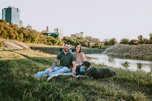 Couple relaxing on grassy riverbank with their dog, city skyline visible in the golden background.
