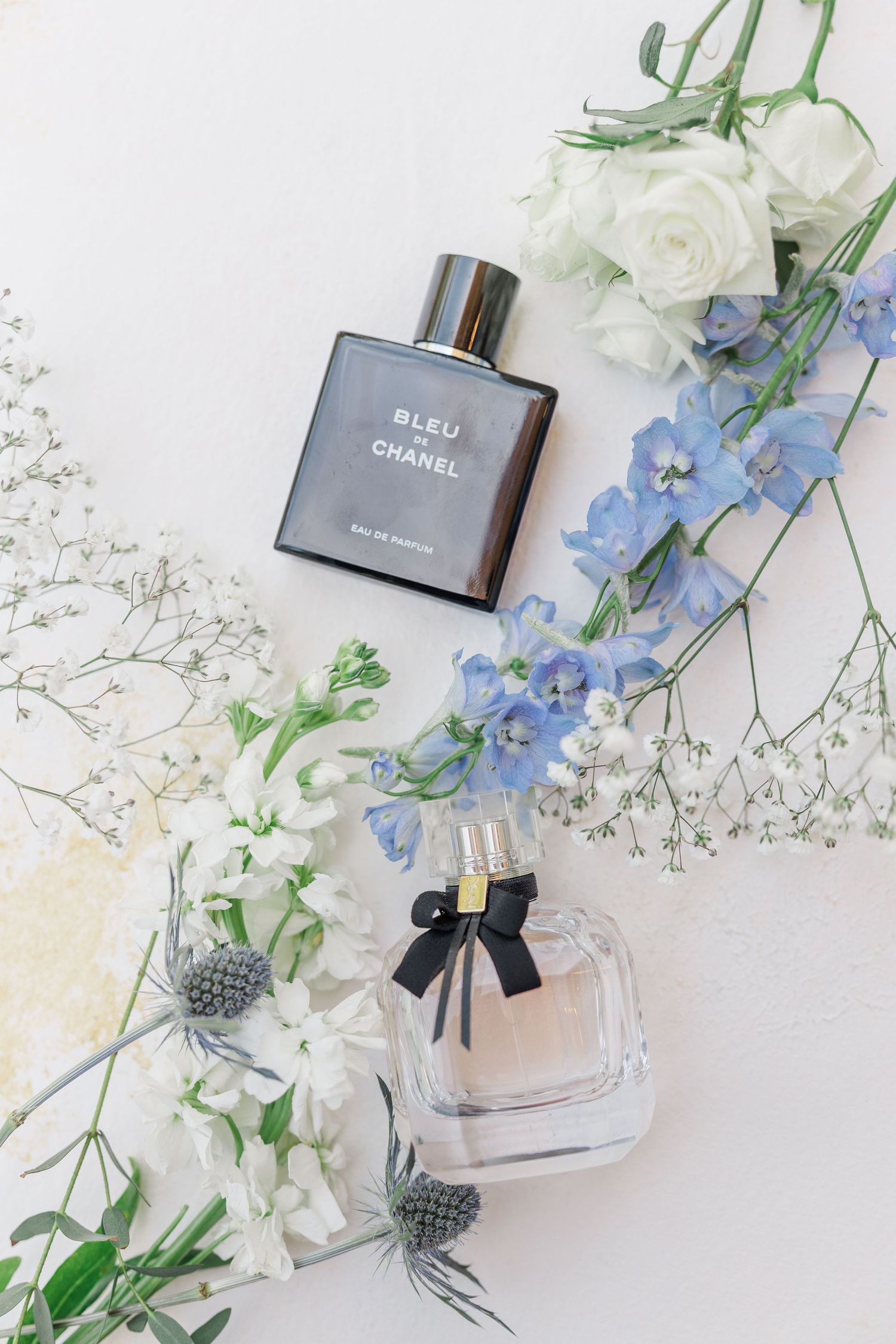 Bleu de Chanel and a floral perfume bottle surrounded by white roses, blue delphiniums, and baby's breath on white.