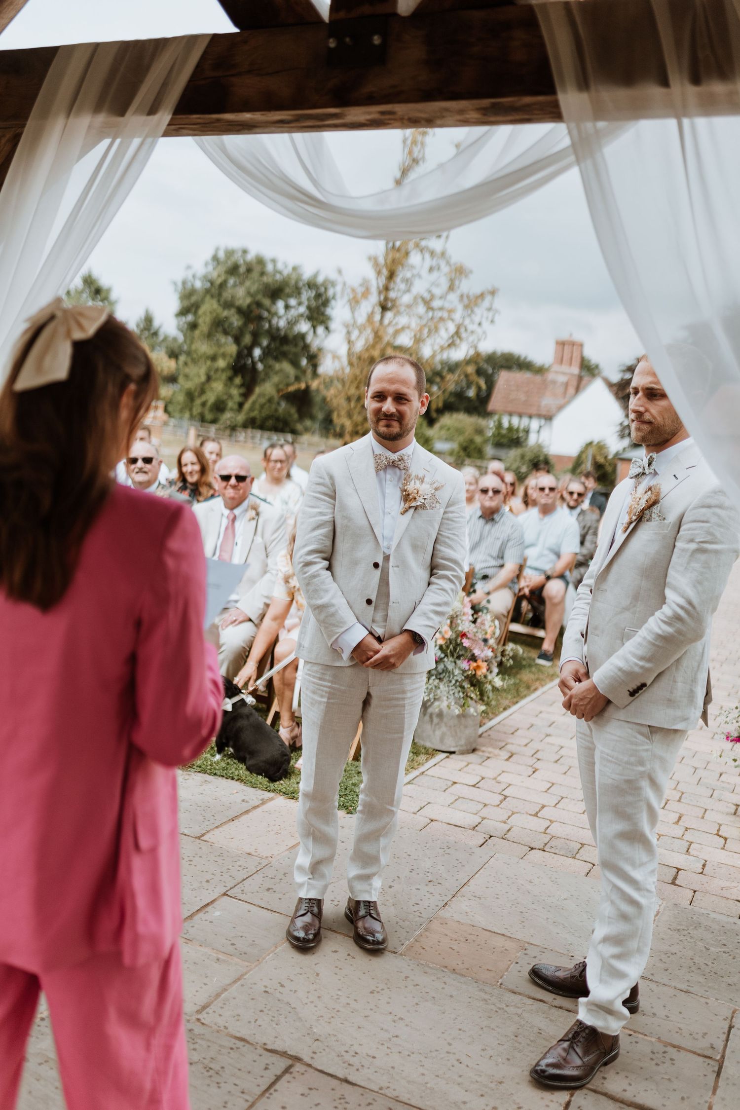 Groom in light gray suit awaits bride at outdoor wedding ceremony, with guests seated behind.