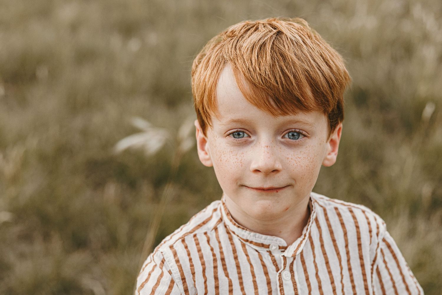 Red-haired boy with blue eyes smiling softly in a field, wearing a striped shirt in a warm outdoor portrait.