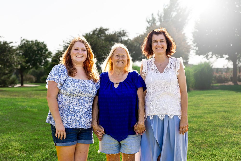 Three women smiling together outdoors on green grass with sunlit trees in the background.