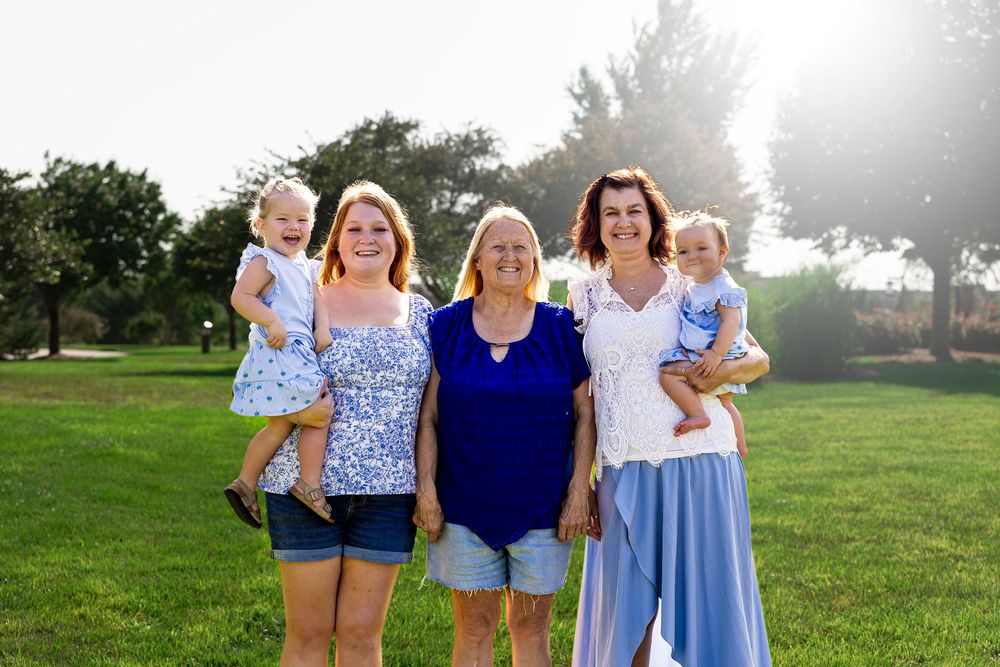 Three generations of women pose outdoors on green grass, two holding toddlers, smiling in a sunny park setting.