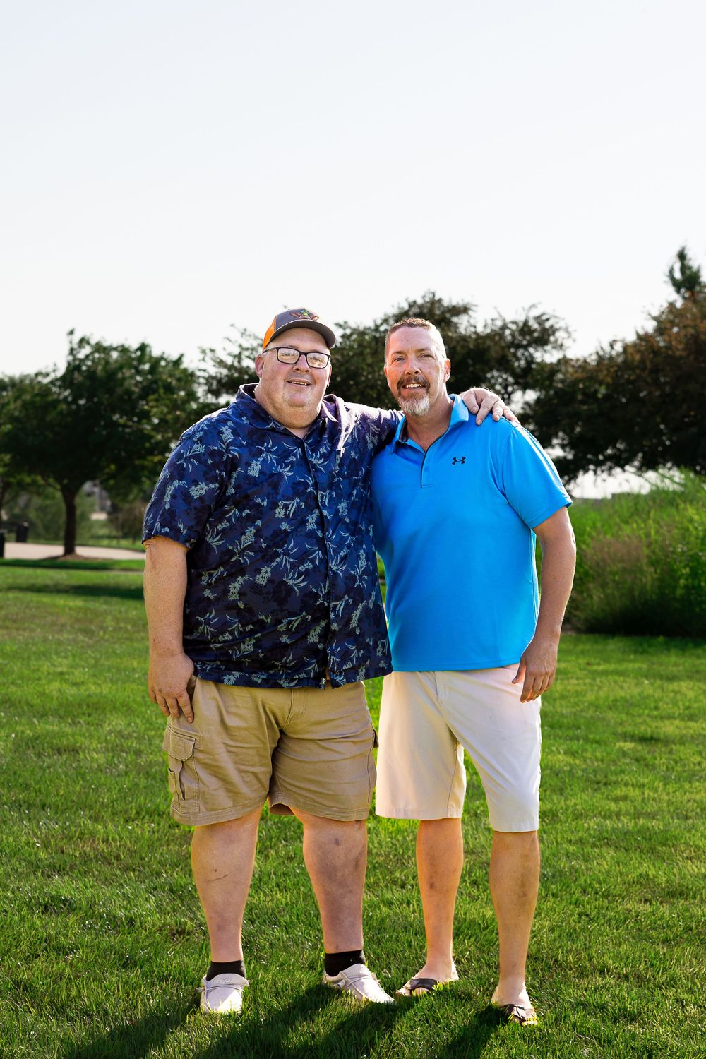 Two smiling men pose together on a green golf course lawn on a sunny day.
