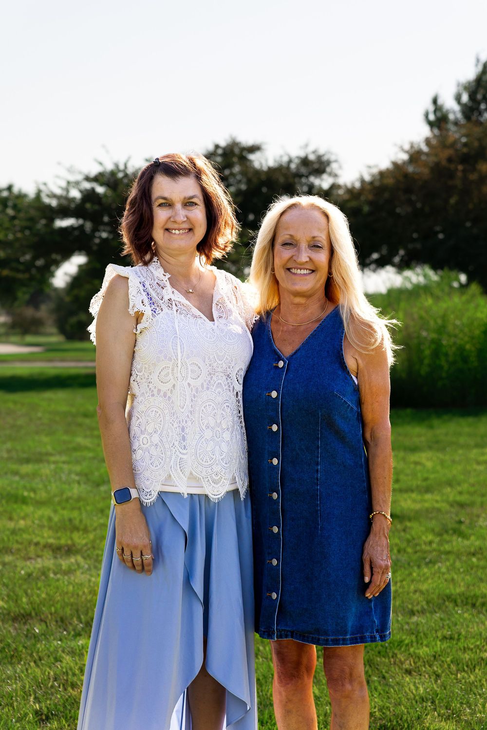 Two smiling women in summer outfits pose together on a green lawn with trees in the background.