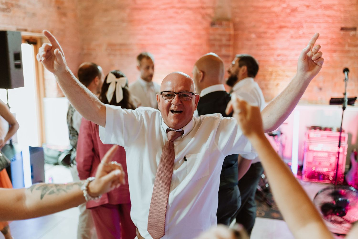 Elderly man joyfully dancing with arms raised at a wedding reception in a brick-walled venue.
