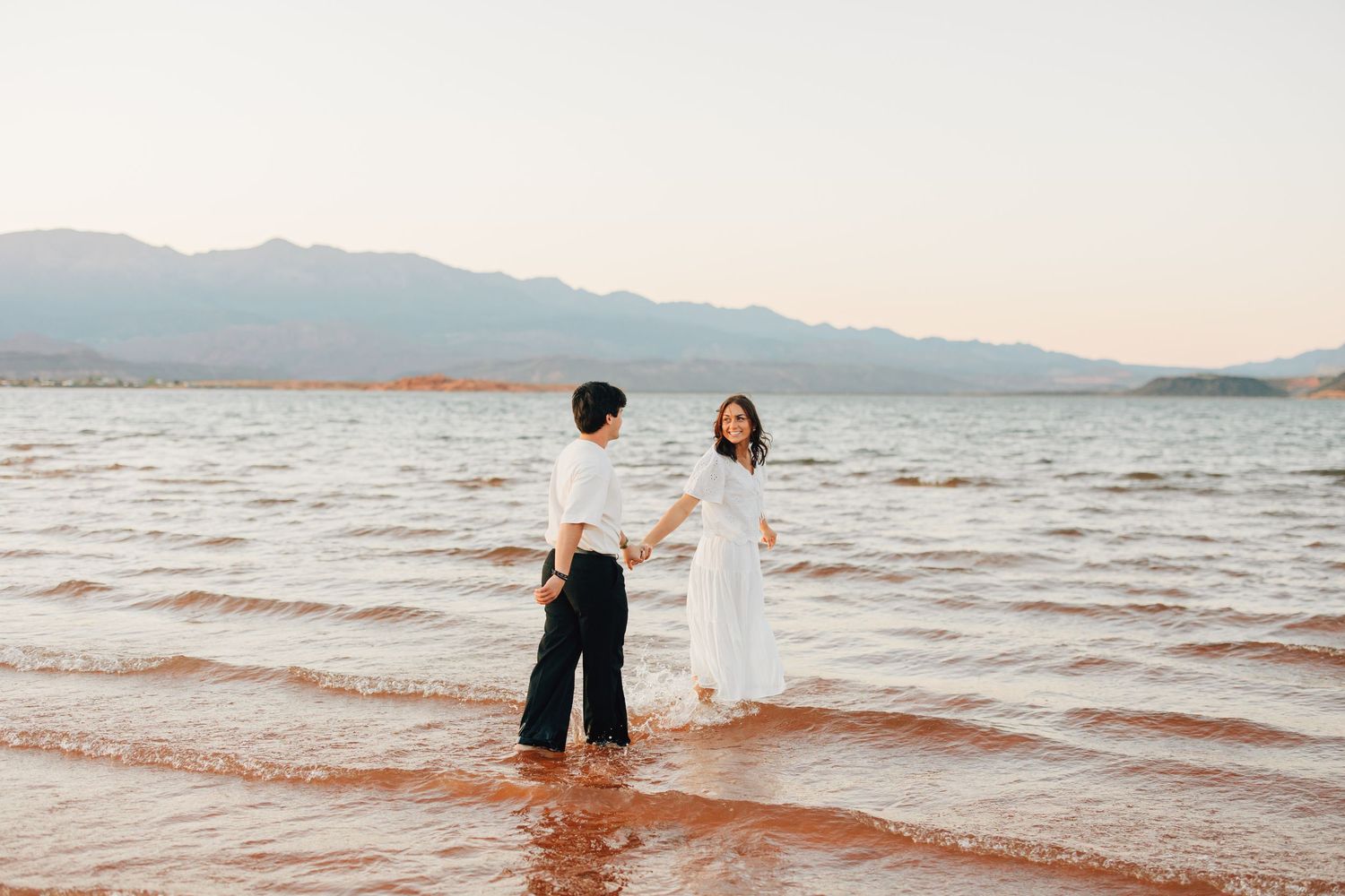 Couple holding hands wading in shallow ocean water at sunset with mountains in the background.