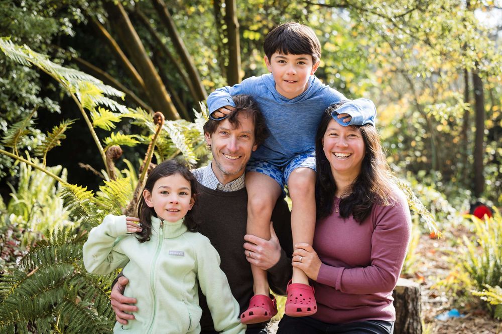 Happy family of four posing outdoors in a lush green forest setting, smiling together on a sunny day.