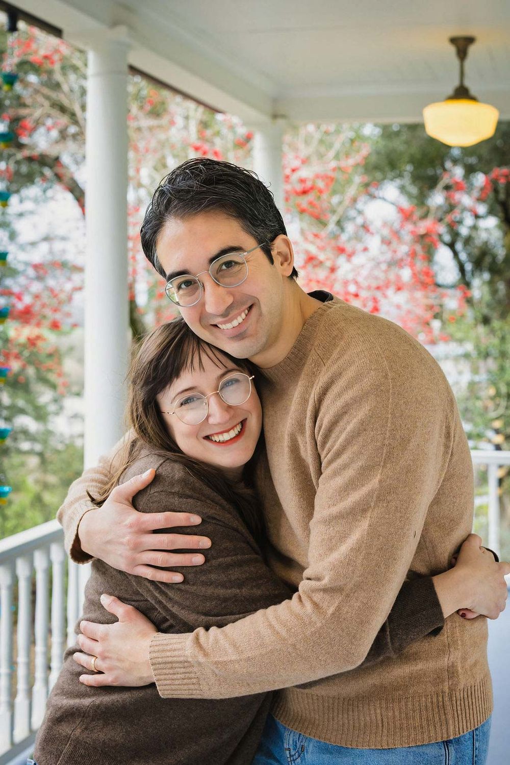 Smiling couple in glasses hugging on a porch with red flowers, wearing cozy sweaters in autumn.