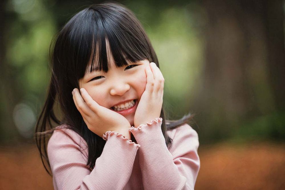 Smiling Asian girl with bangs resting her face in her hands outdoors in a park setting.