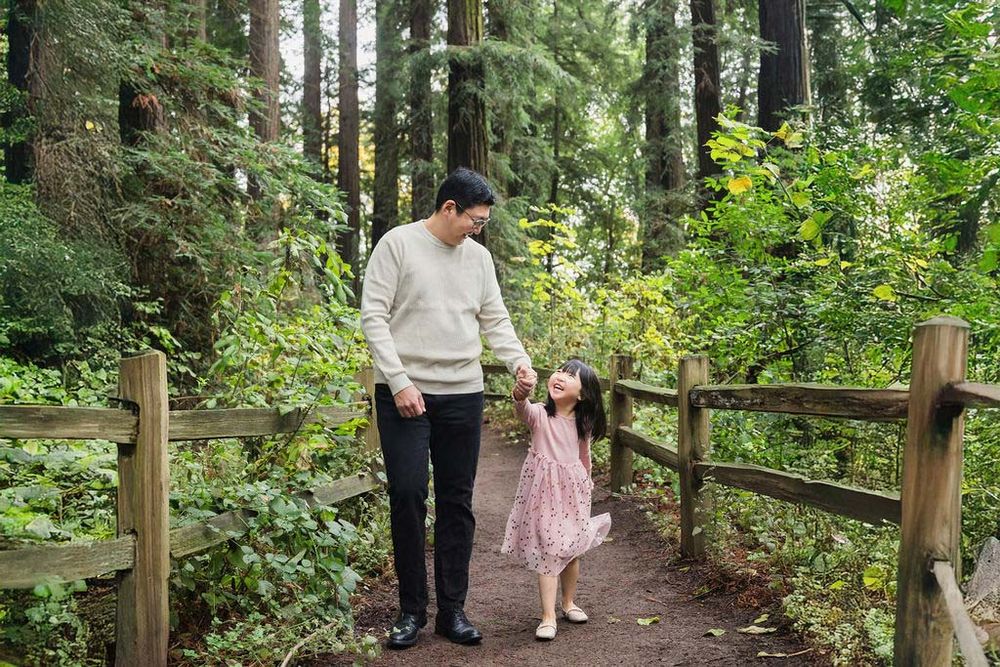A father and young daughter hold hands on a wooden forest path surrounded by tall redwood trees and lush greenery.