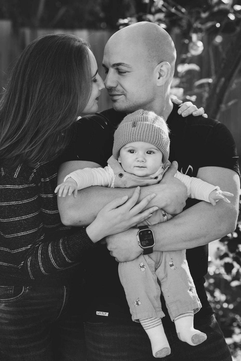 Black and white photo of a loving couple holding their smiling baby outdoors, family portrait.