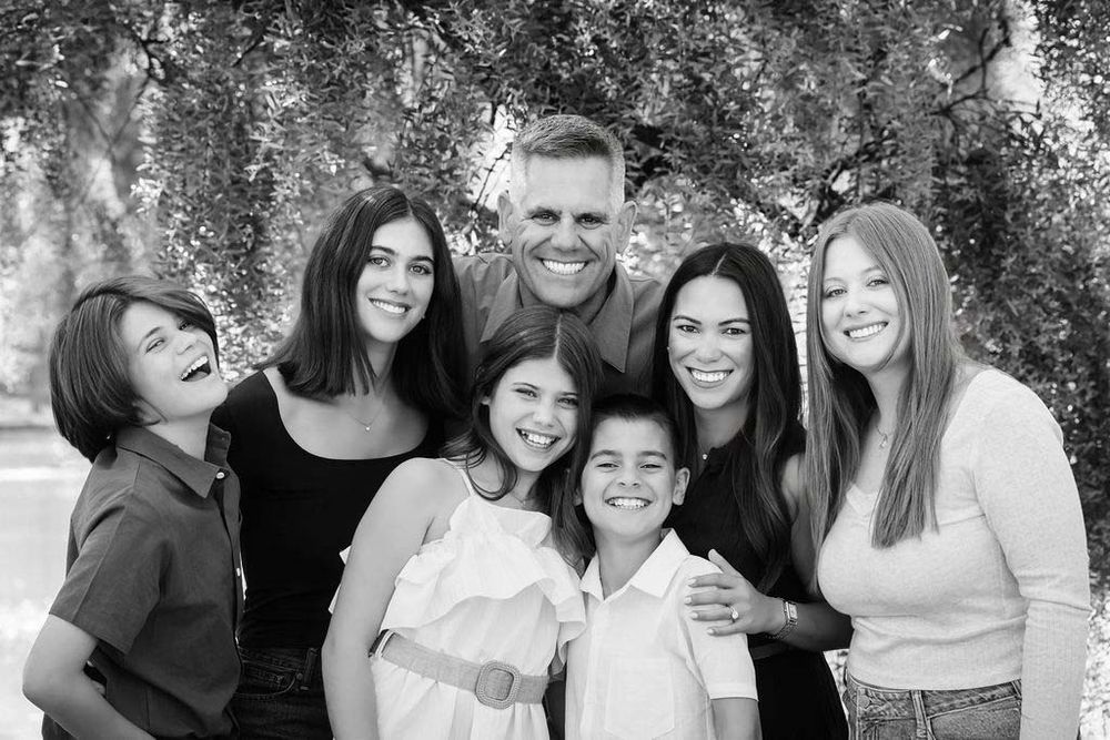 Black and white family portrait of parents with four children smiling outdoors under a weeping willow tree.