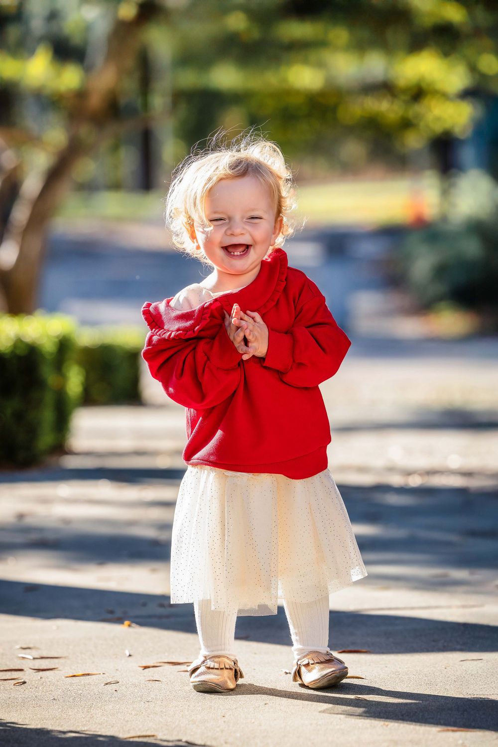 Happy toddler girl in red sweater and white tutu skirt laughing joyfully in sunny park.