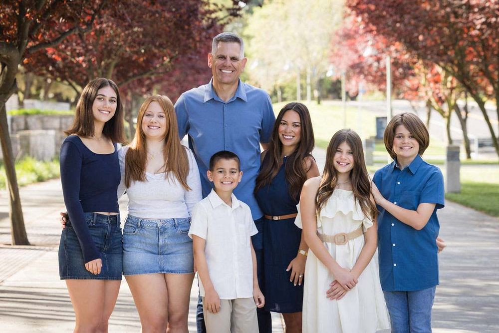 A smiling family of seven poses outdoors on a sunny day, surrounded by colorful autumn trees on a paved path.
