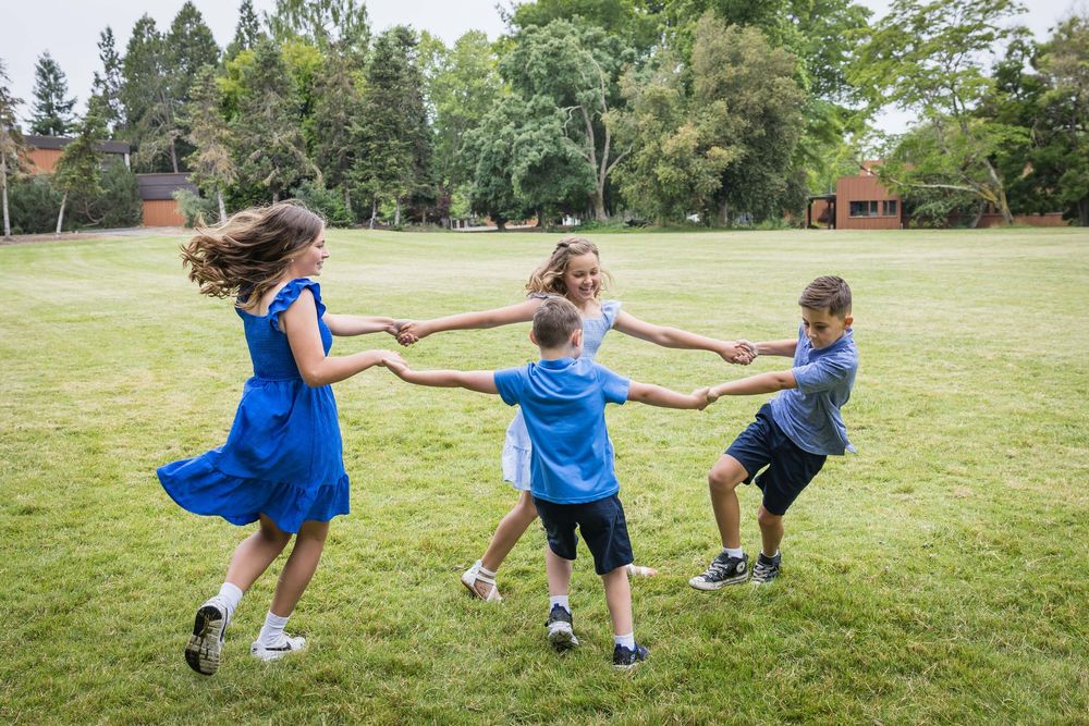 Four children holding hands and spinning in a circle while playing outdoors on a green grass field.