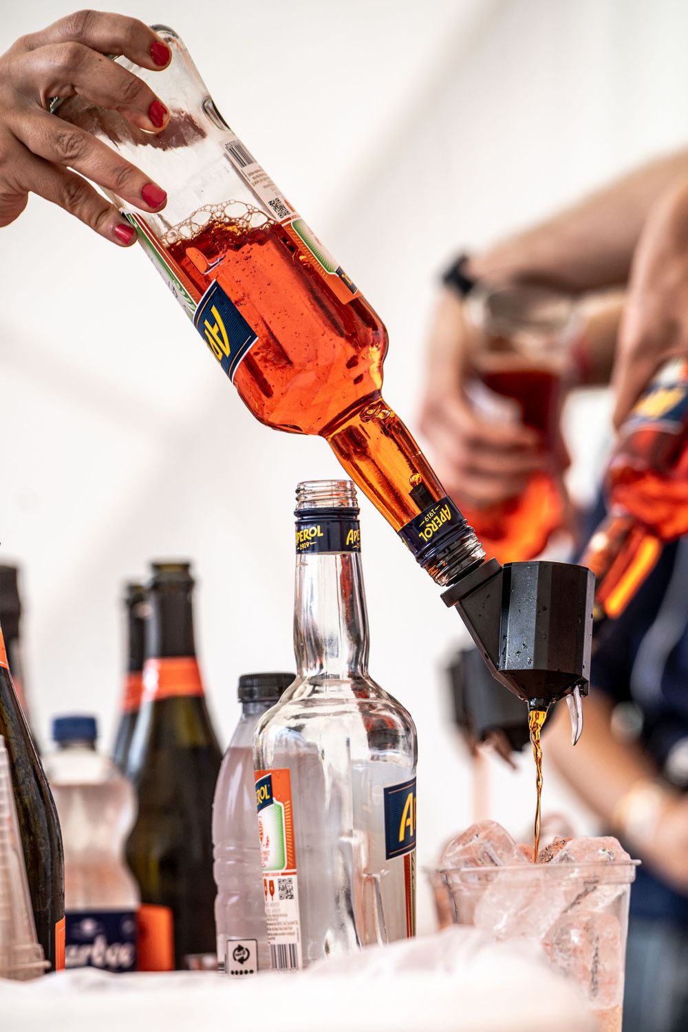 Bartender pouring Aperol spirits into glasses with ice, surrounded by bottles at a bar setup.