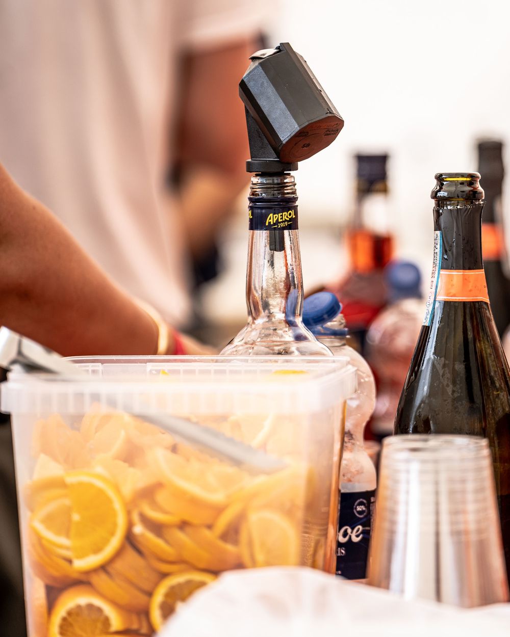 Bar setup with Aperol bottle, sliced oranges in a pitcher, sparkling wine, and plastic cups for cocktail service.