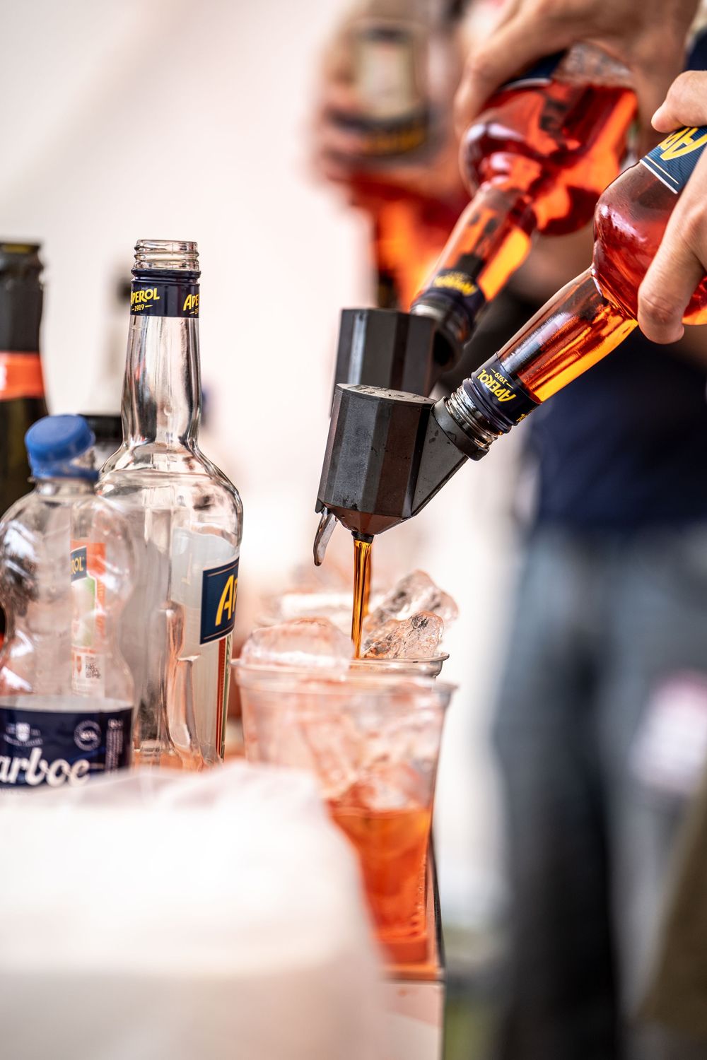 Bartender pouring dark liquor into plastic cups with ice, surrounded by bottles at a bar event.