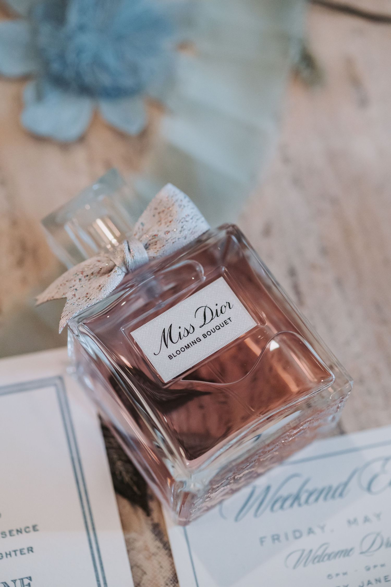 Miss Dior Blooming Bouquet perfume bottle rests beside wedding weekend itinerary cards and blue flowers.